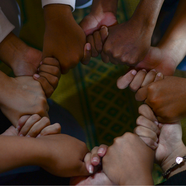 Overhead close-up of several people's hands stacked together in a fist-bump circle, forming a unity gesture over a green and yellow patterned mat.